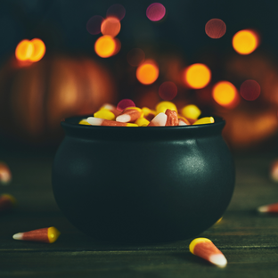 Small black cauldron shaped pot in the forefront, filled with candy corn with a few pieces on the surface the cauldron is sitting on. Blurred orange and yellow circles and shapes in the indistinct background.