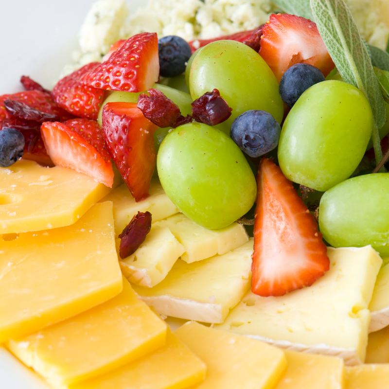 Close view of a plate with slices of cheese, strawberries, green grapes, dried cranberries, and blueberries mixed together.