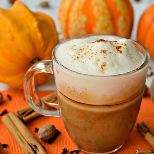 Coffee drink in glass mug with handle with steamed milk on top, pumpkins in the background, cinnamon sticks and spices on the surface surrounding the mug