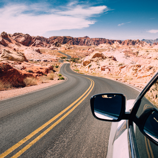 View out the side of a car rear window, facing forward. Rearview mirror in view, with winding black road with double yellow line down the center. Desert rock formations along the road, and blue sky with some wispy white clouds.